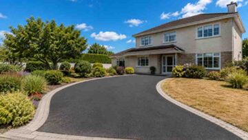 Newly completed smooth black tarmac driveway with light granite block paving borders leading to a detached 1970s Irish family home in County Kildare, with a straw-coloured summer lawn, mature shrubs and bright blue sky.